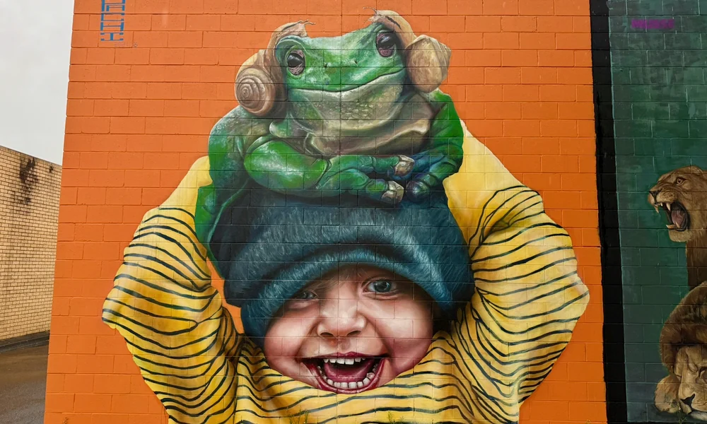 A mural of a smiling child holding a large green frog above their head against an orange wall.