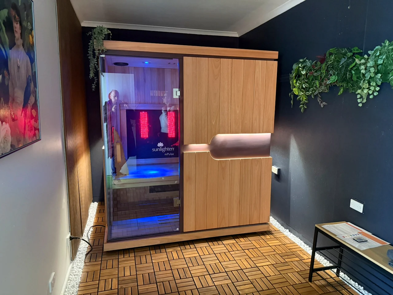 An infrared sauna in a dark-walled room, surrounded by timber flooring, white stones and trailing plants.