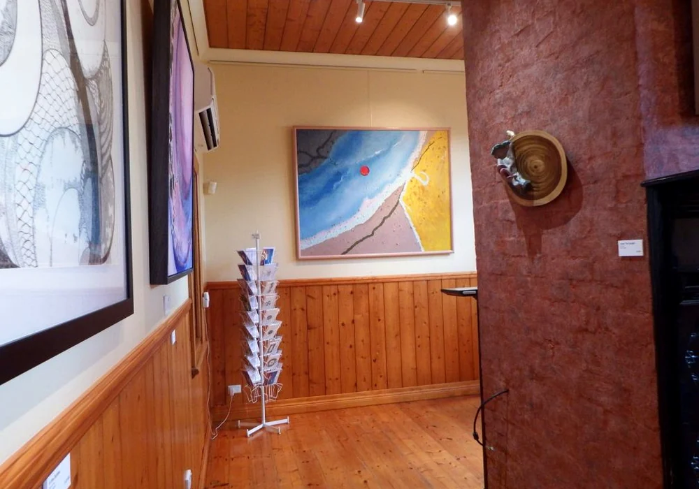 A view through Matchbox Gallery past framed artworks, timber panelling, a display rack and a textured red wall.