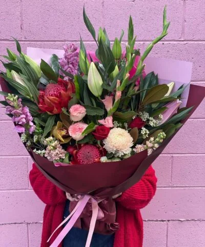 A large bouquet of pink, burgundy, cream and green flowers held against a pink brick wall.