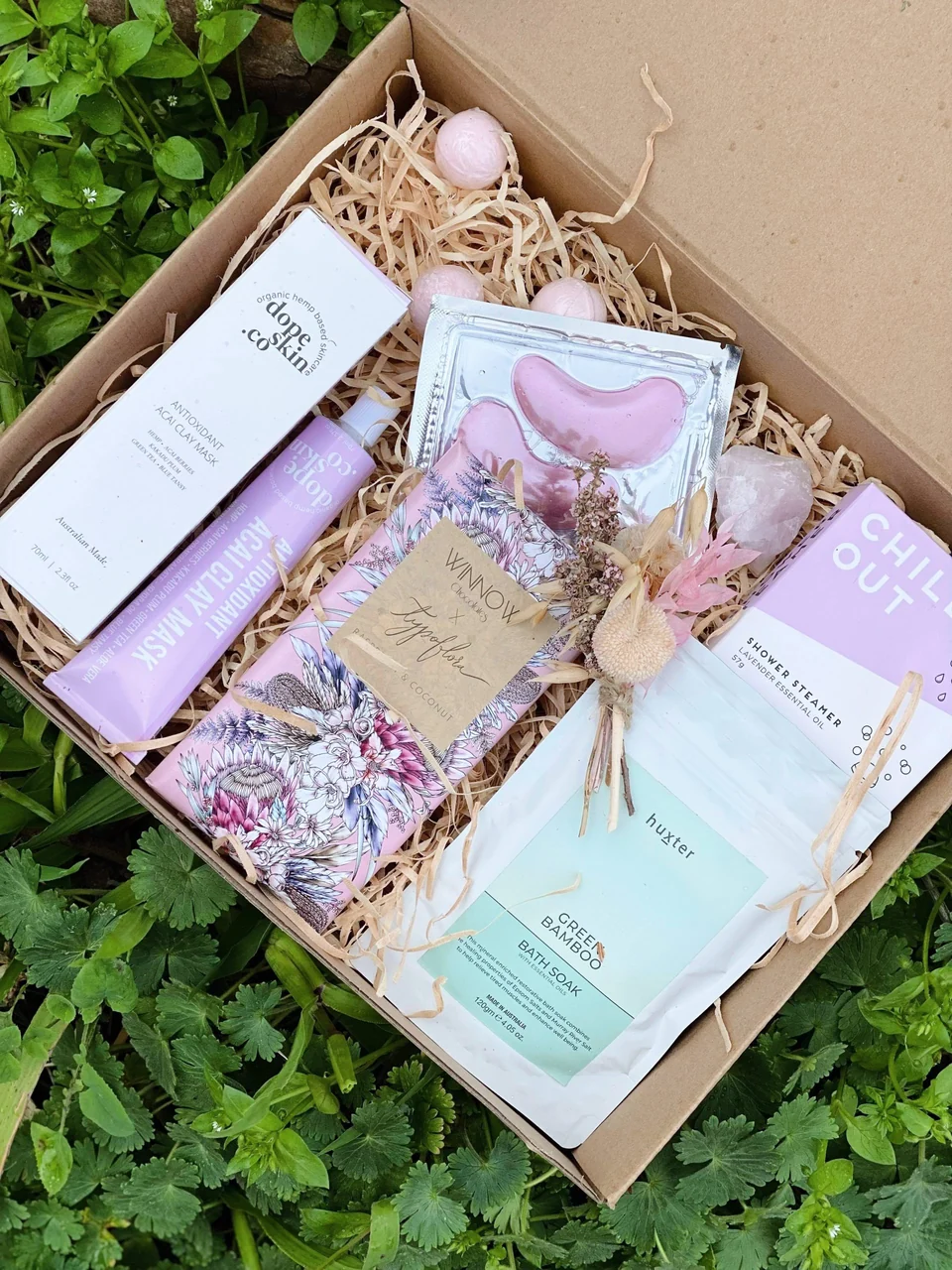 A gift box of pink self-care items, chocolate and dried flowers rests among green groundcover.