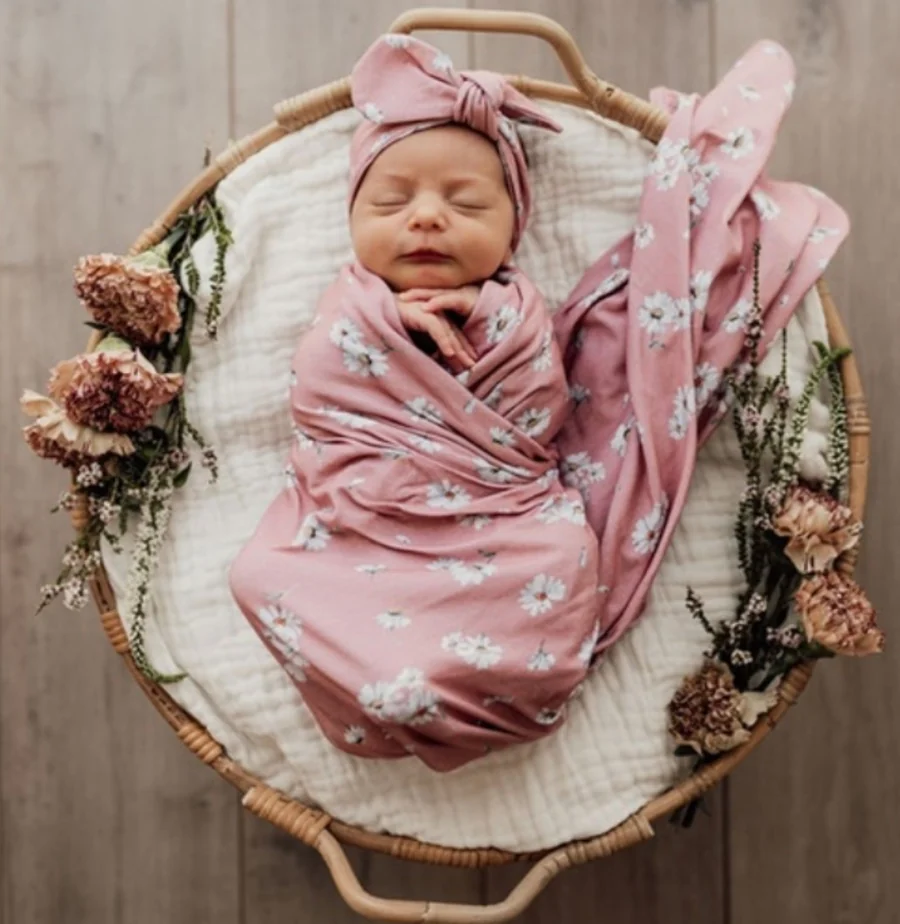 A newborn wrapped in pink floral fabric lies in a woven basket beside dried flowers.
