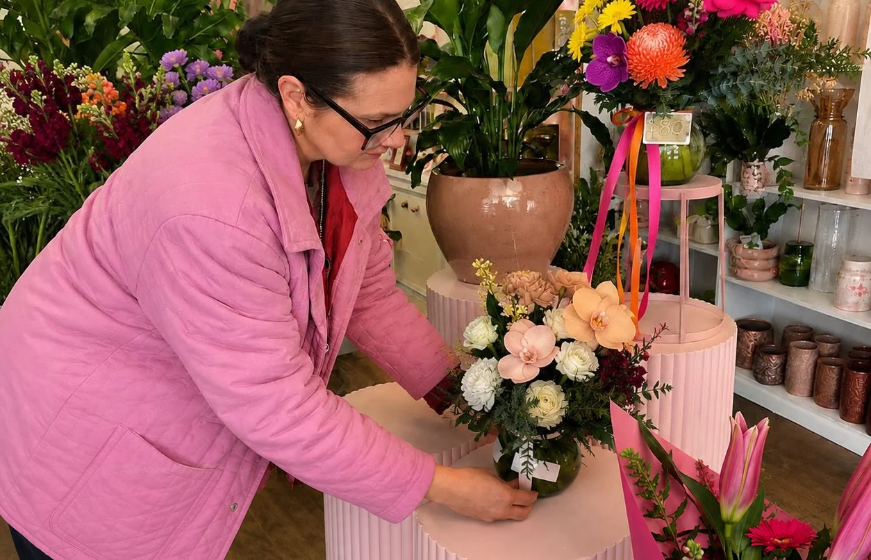 A florist in a pink jacket arranges peach and cream flowers inside a shop filled with bright flowers, plants and pink vessels.