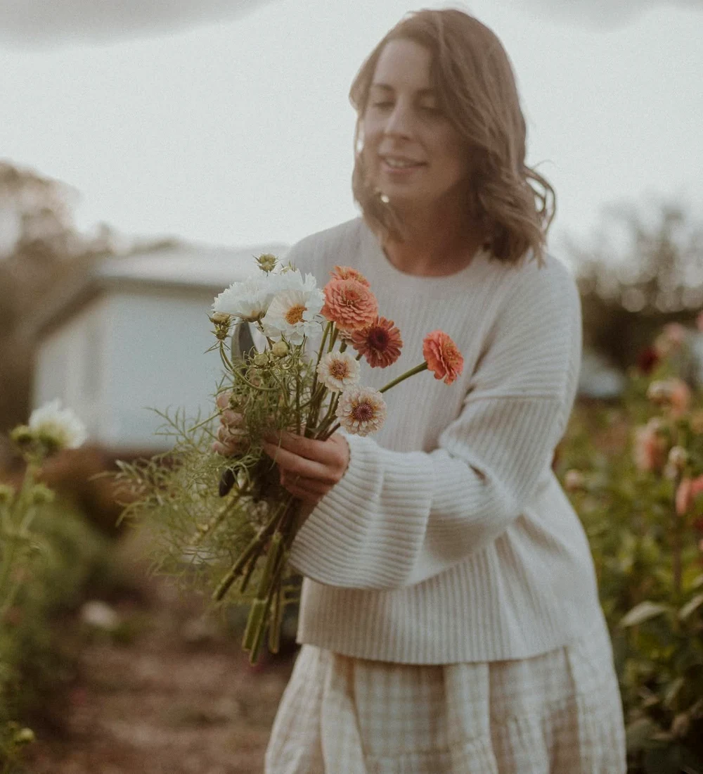 A woman holding freshly cut pink and white flowers among garden beds.