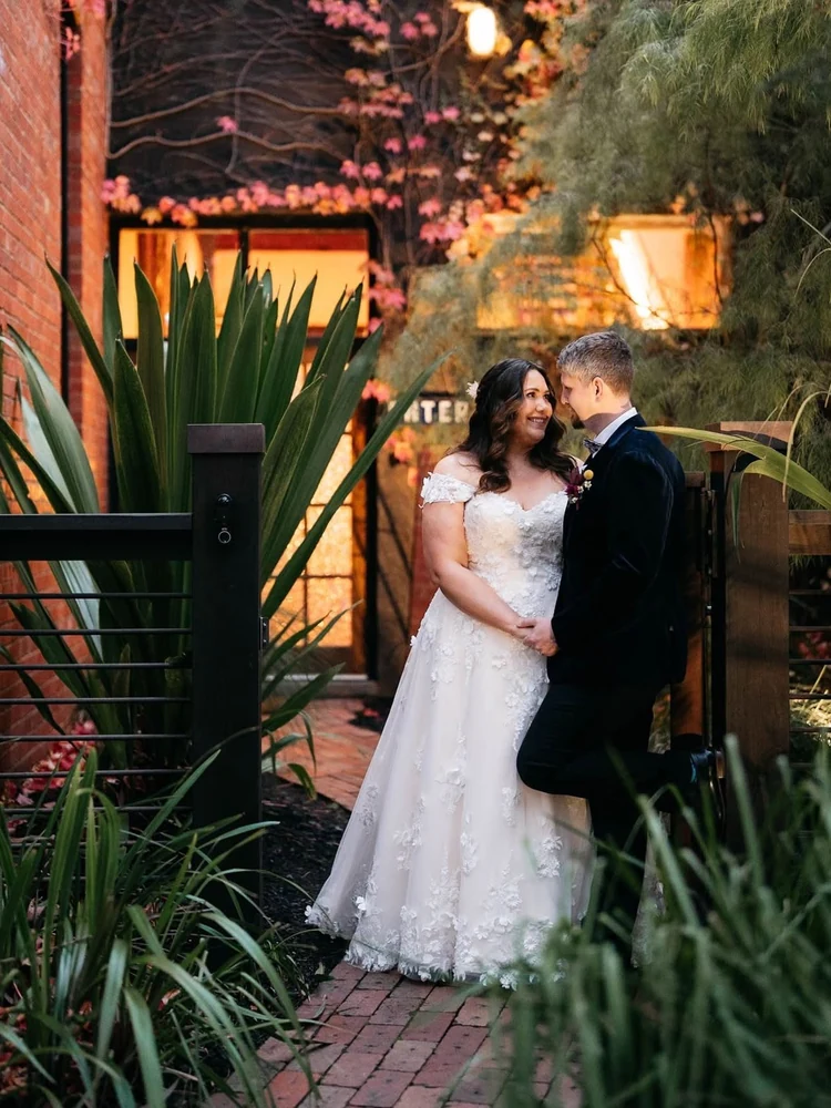 A newlywed couple stand together in a garden courtyard surrounded by foliage and warm light.