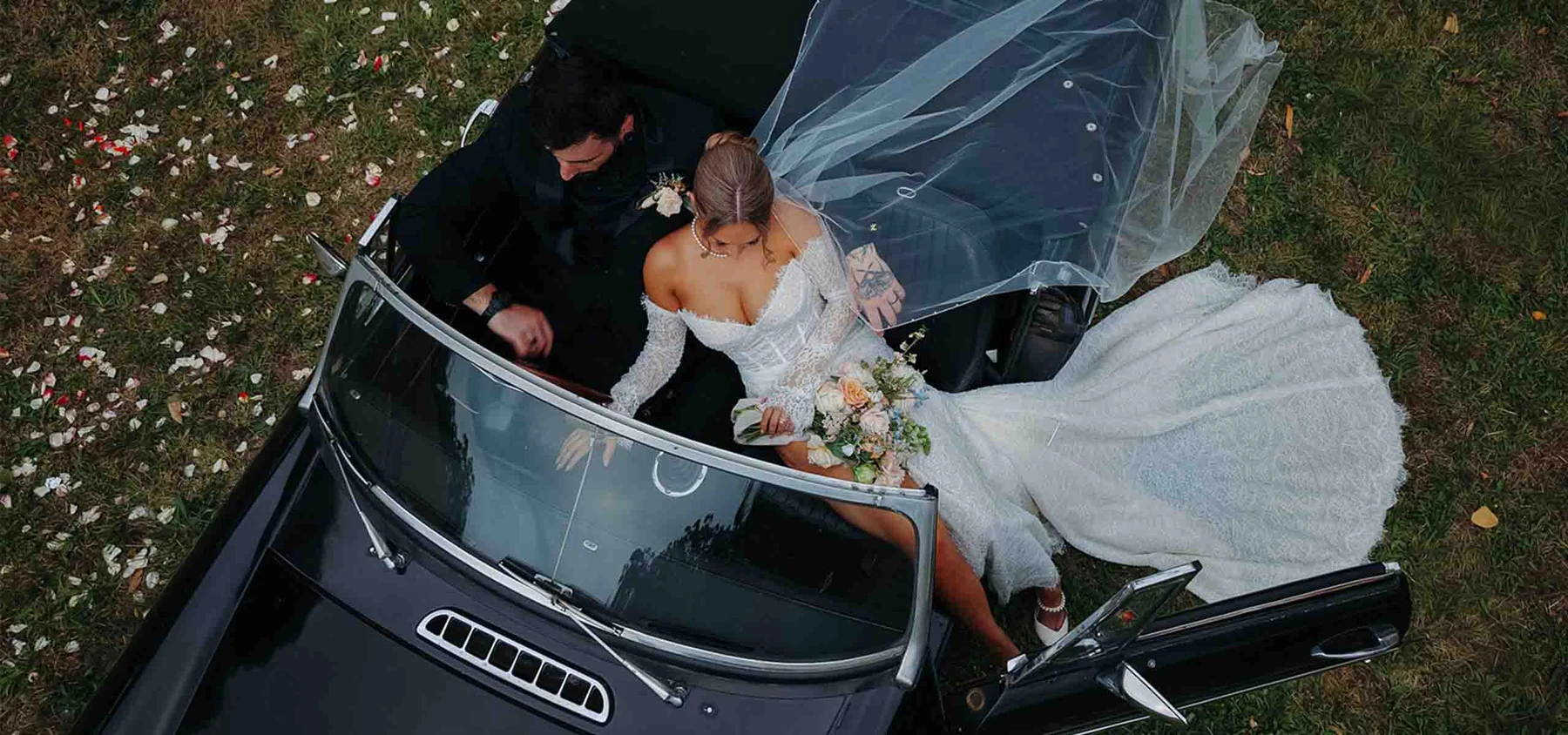 Overhead view of newlyweds seated in a black convertible surrounded by flower petals.