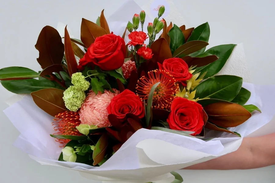 A bouquet of red roses, pink chrysanthemums and native flowers held against a pale background.