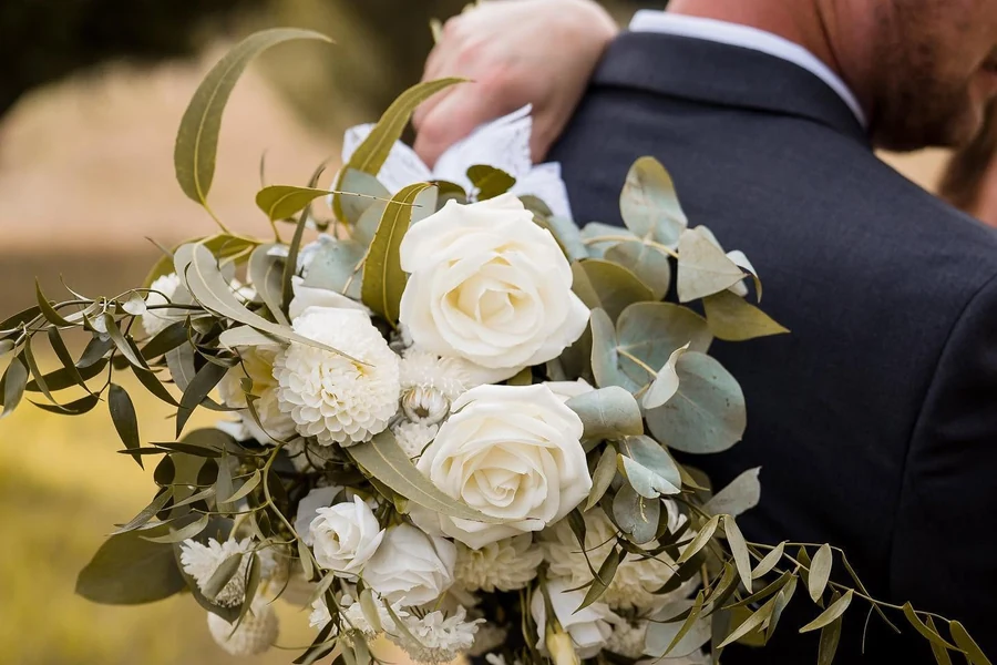 A couple embrace outdoors with a cascading bouquet of white roses and greenery in the foreground.