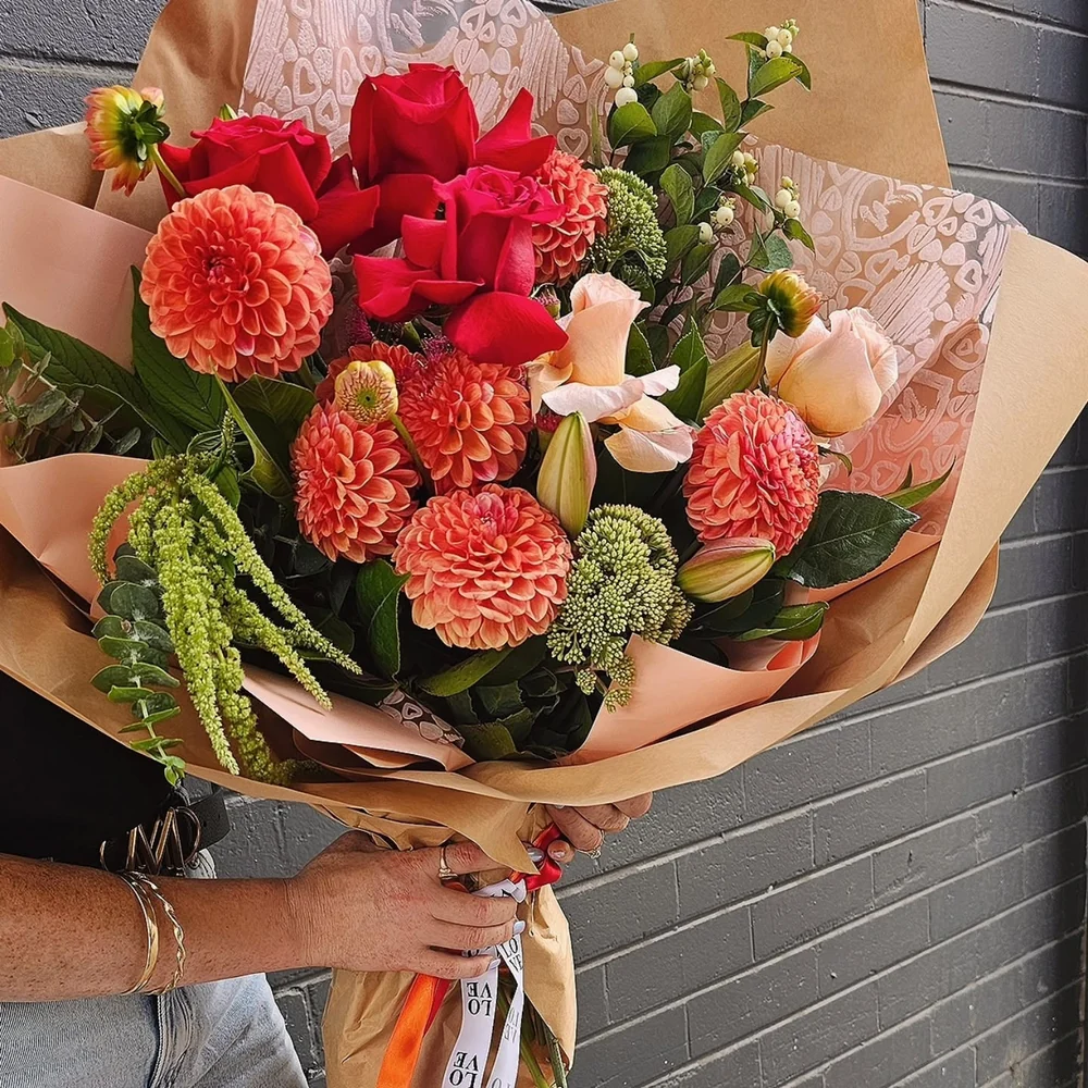 A large bouquet of red roses, peach dahlias, lilies and green foliage held against a dark brick wall.