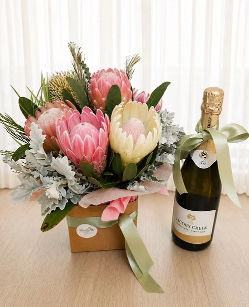 Pink and cream proteas arranged beside a ribboned bottle in a bright gift presentation.