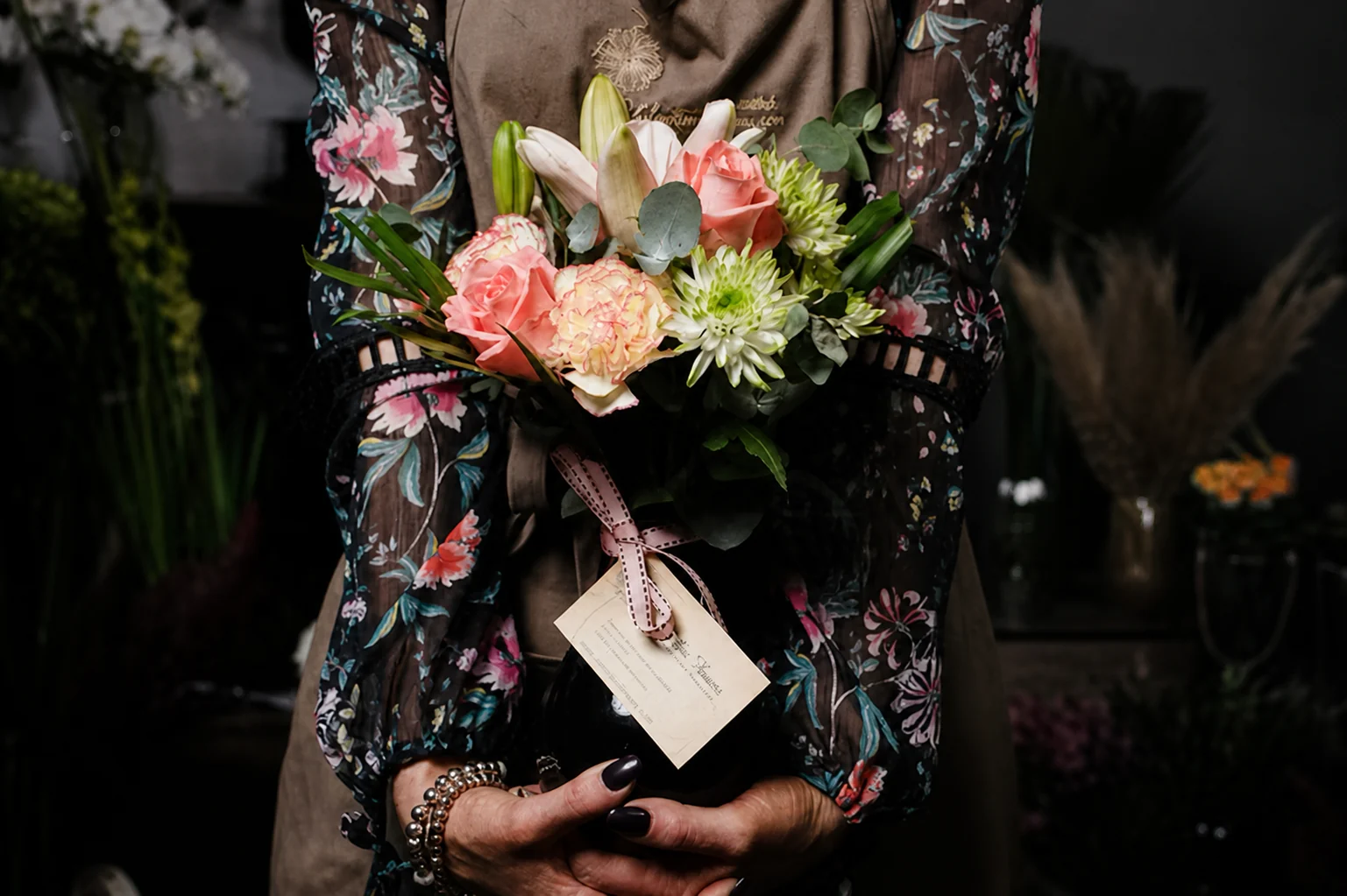 A person in a floral jacket holds a pink, cream and green bouquet against a dark background.