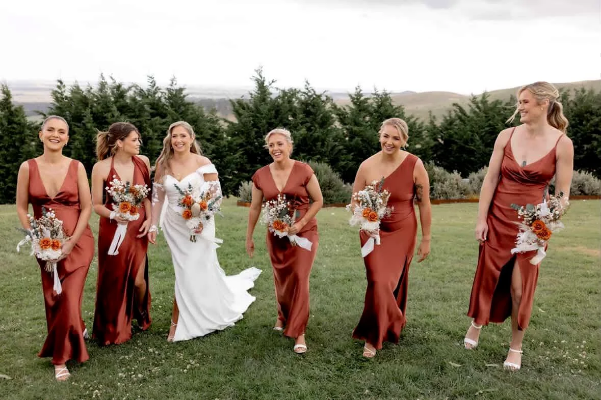 A bride and five attendants walk outdoors carrying coordinated rust, white and green bouquets.