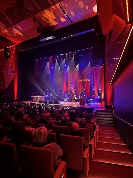 An audience faces a concert stage illuminated in red and violet inside the Gippsland Performing Arts Centre auditorium.