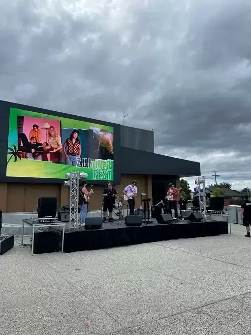 Musicians perform on an outdoor stage beside Gippsland Performing Arts Centre beneath a cloudy sky.