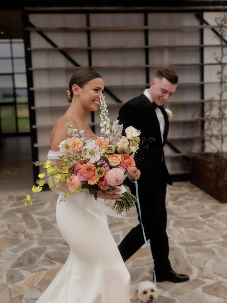 A smiling bride holds a loose bouquet of peach, pink, white and yellow flowers as the groom walks behind her.