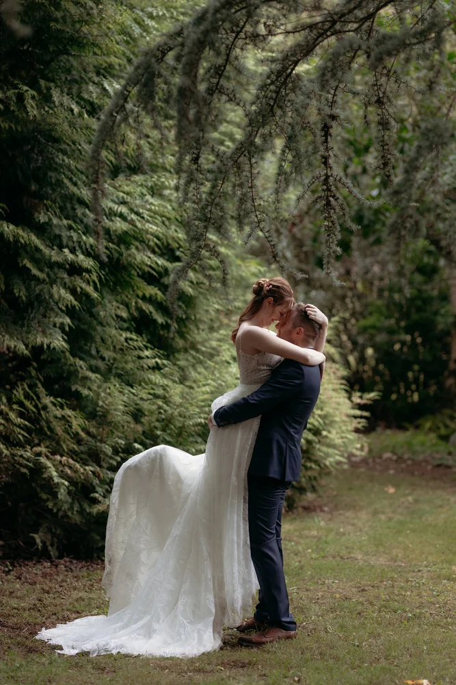 A newlywed couple embrace beneath ferns in a forest clearing.