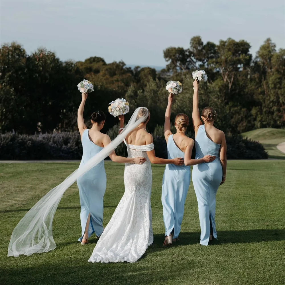 A bride and three attendants in pale blue dresses raise their bouquets on a lawn.