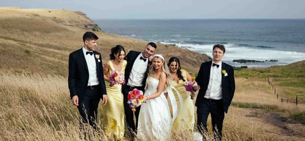 A wedding party in black suits and pale yellow dresses walks through a coastal landscape.