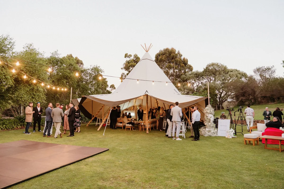 Wedding guests gather around a lit canvas marquee on a lawn at dusk.