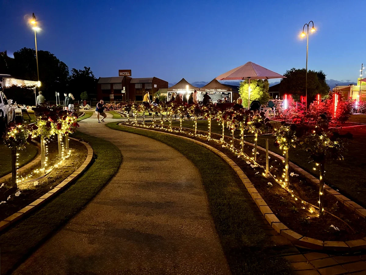 People move through illuminated garden beds and community stalls in Morwell at dusk.