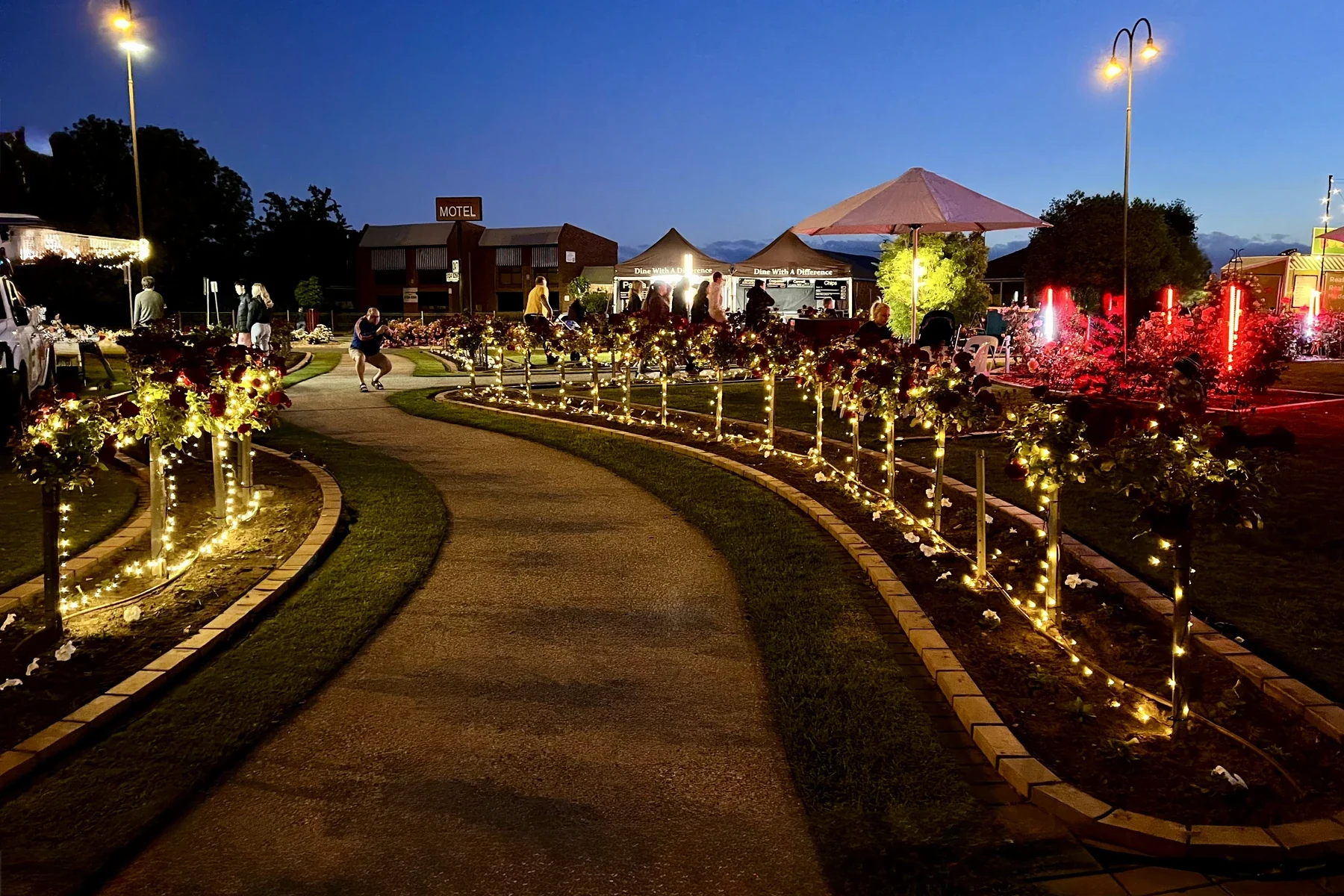 People move through illuminated garden beds and community stalls in Morwell at dusk.