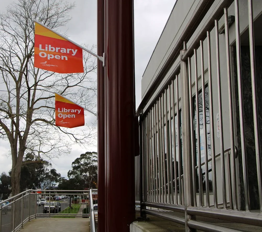 Two orange Library Open flags project from the Morwell Library façade on an overcast day.